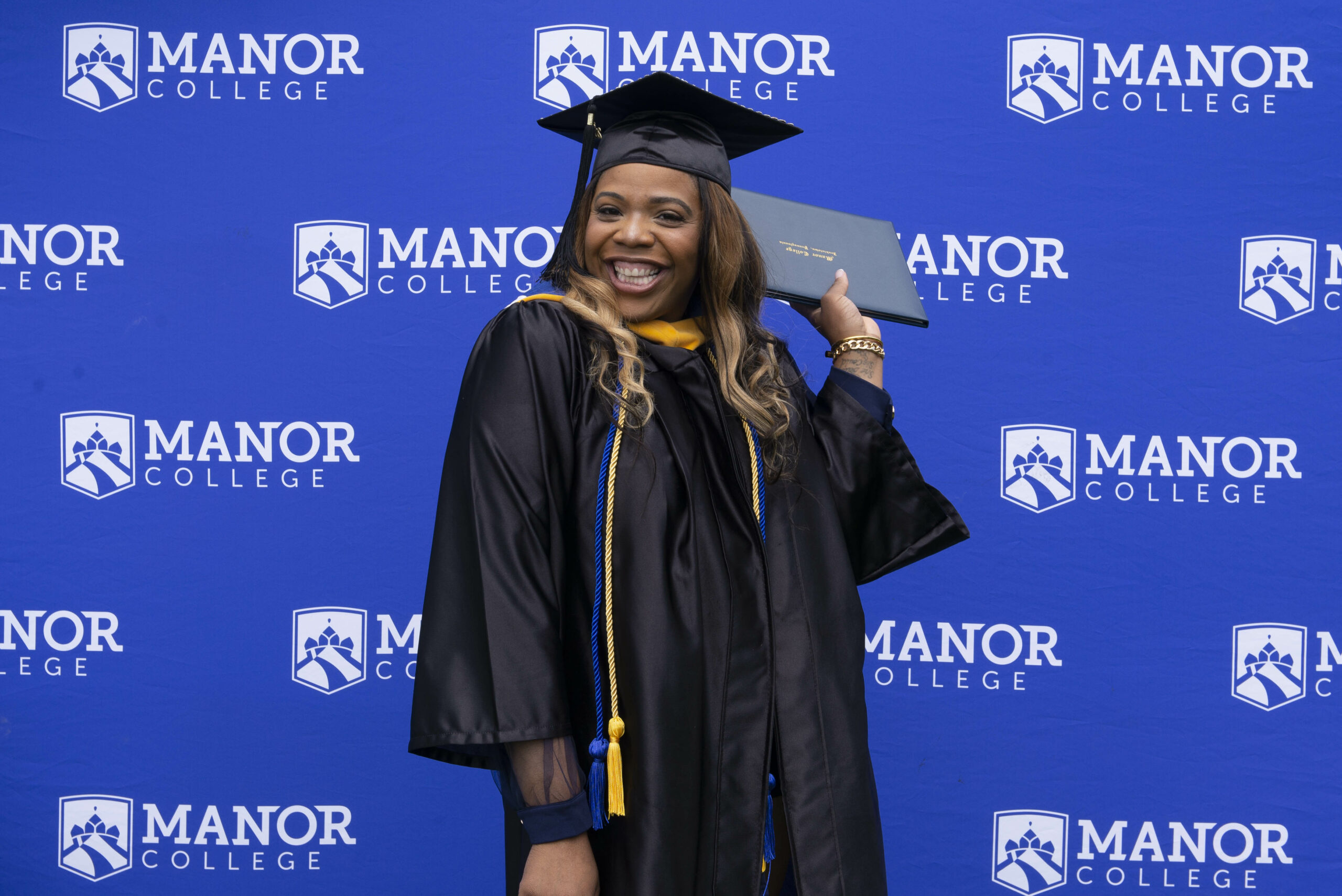 A woman stands in graduation clothing in front of a Manor College background