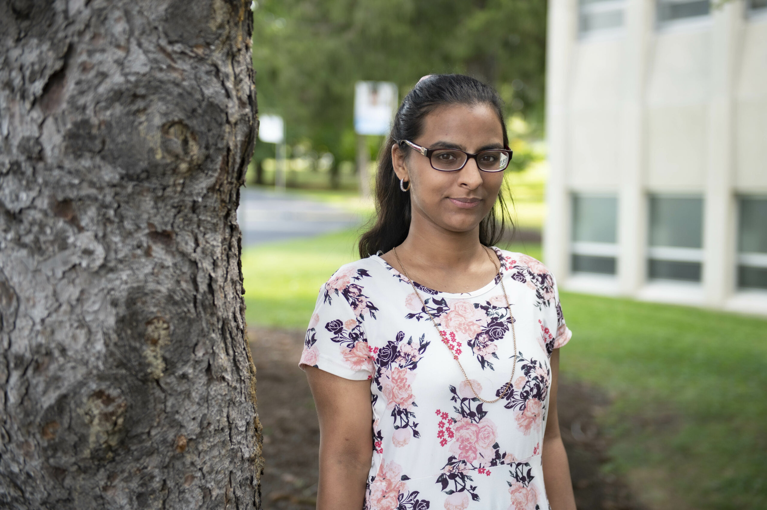 A woman in a white blouse with a floral pattern stands next to a tree.