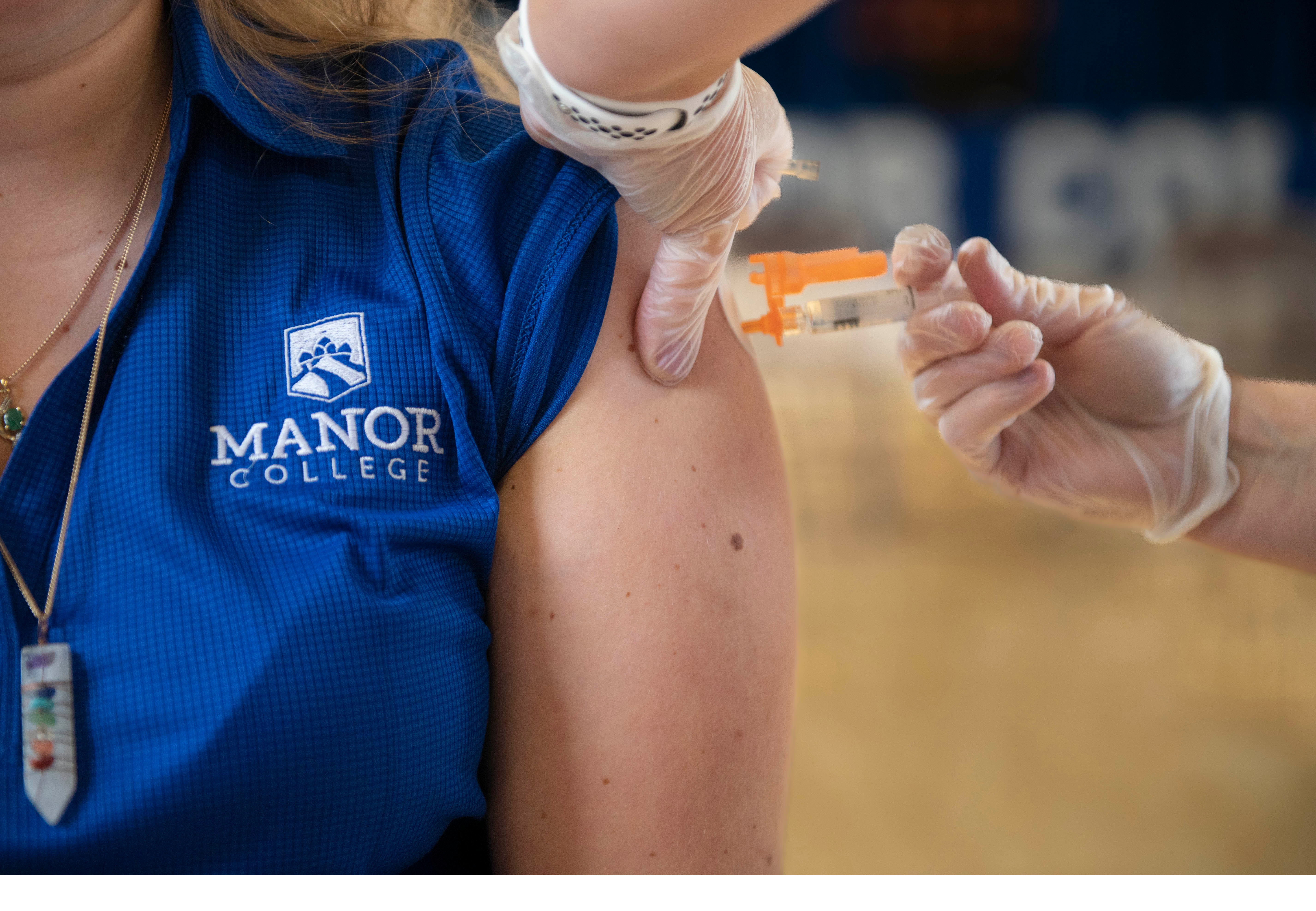 Close-up shot of student wearing Manor college shirt getting vaccine from provider
