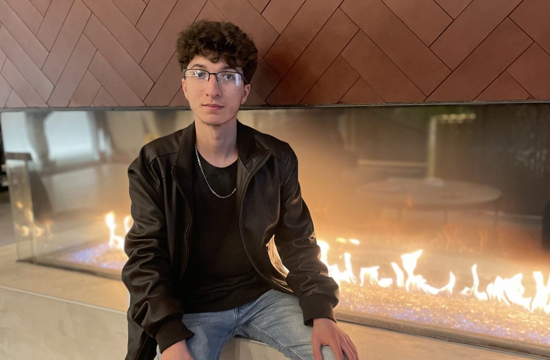 A student in a leather jacket sits next to a fireplace. 