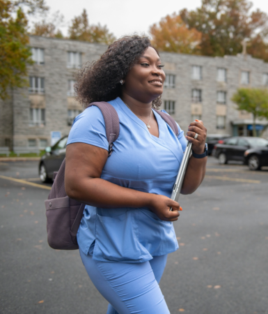 Manor College student in scrubs walking across the parking lot with a backpack and books.