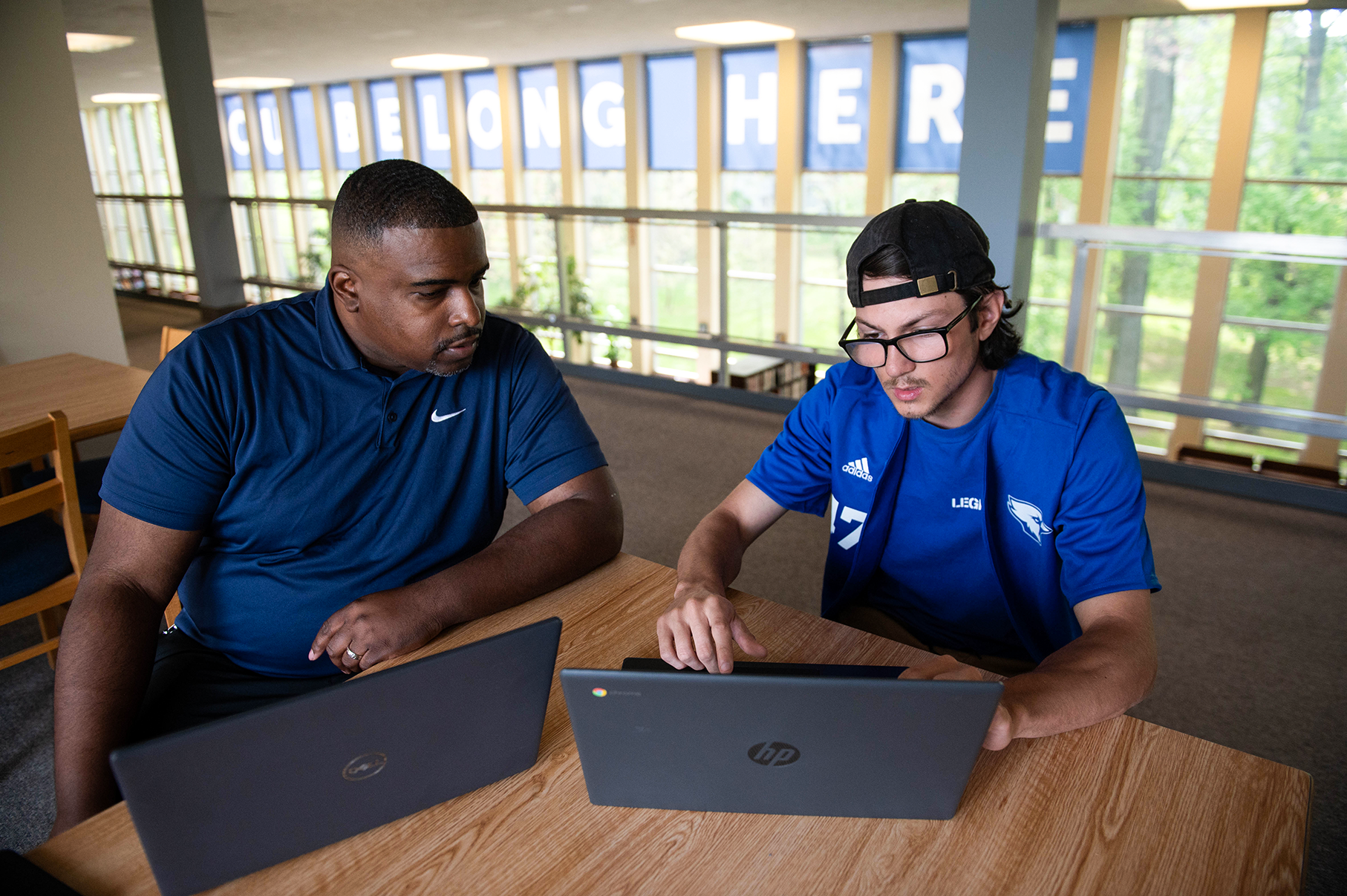 A Manor College student in a cap and glasses works on a laptop alongside a professor in the library.